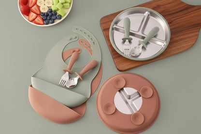 Ensemble repas bébé en inox et silicone – assiette compartimentée, bol amovible, cuillère, fourchette et bavoir en silicone.